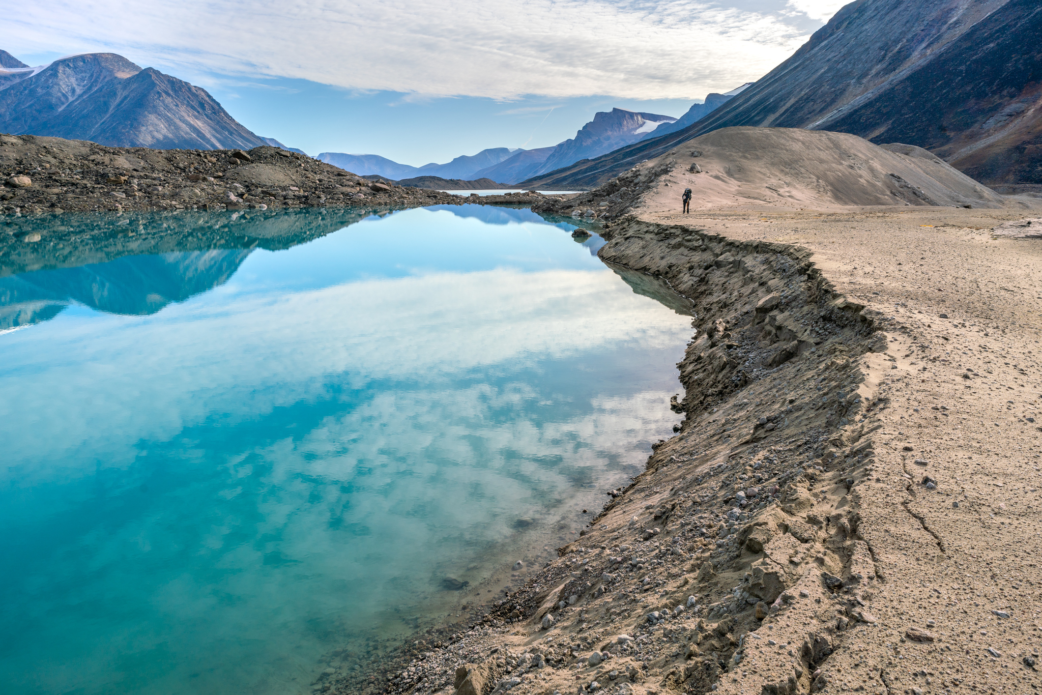 Hiker walking by a lake through remote arctic valley on a partly cloudy summer day. Dramatic arctic landscape of Akshayuk Pass, Baffin Island, Canada. Enormous glacier moraine. Arctic wilderness.