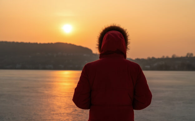 A young man in a red jacket, in the winter on the frozen sea, stands with his back in thought. Against the background of a sunset.